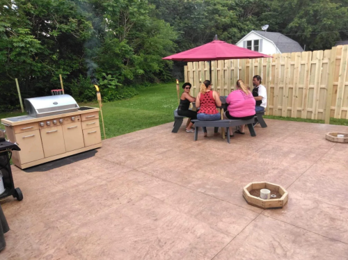 A group of people sitting around a table on a patio, with a grill nearby and a shaded area under a red umbrella.