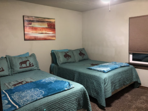 Cozy bedroom with two beds, teal bedding, and a colorful wall art piece. Soft lighting and a window with blinds.