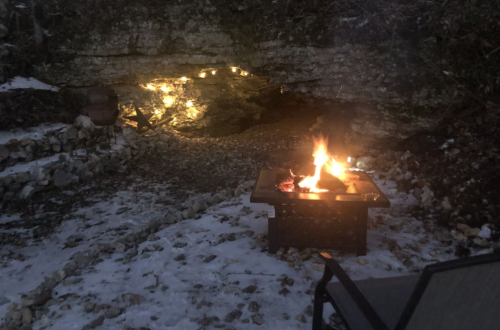 A cozy outdoor scene with a fire pit, snow on the ground, and warm lights illuminating a rocky cave in the background.