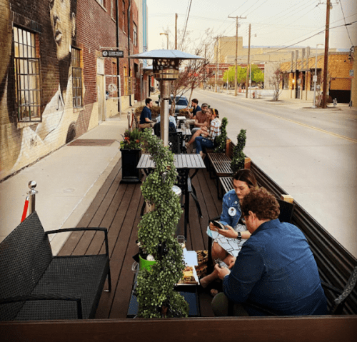 Outdoor dining area with patrons seated at tables, surrounded by greenery and a mural in the background.