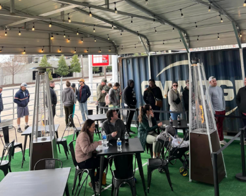 A lively outdoor seating area with people socializing, heaters, and string lights under a canopy.