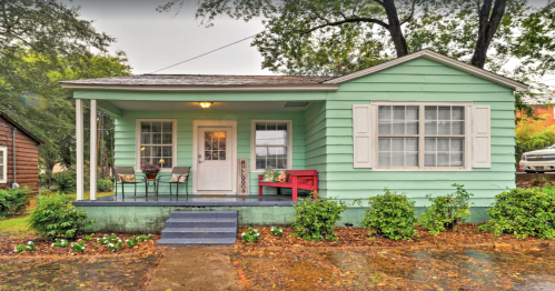 A light green house with a porch, white shutters, and a red bench, surrounded by greenery on a rainy day.
