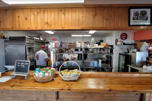 A cozy kitchen with chefs at work, a wooden counter in front displaying snacks and a chalkboard for orders.