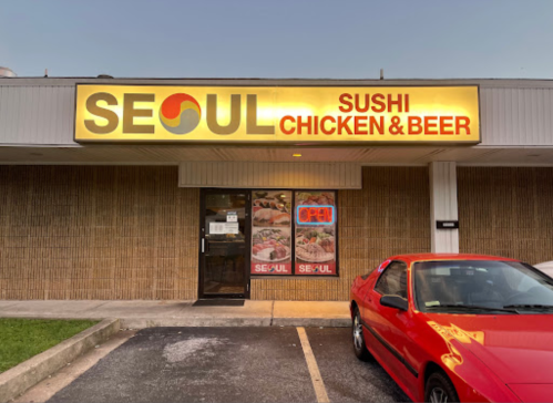 Exterior of a restaurant named "Seoul" featuring a sign for sushi, chicken, and beer, with a red car parked outside.