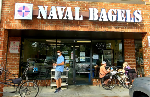 A man stands outside Naval Bagels, with customers seated nearby and bicycles parked in front of the shop.