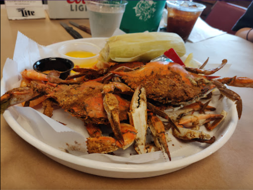 A plate of seasoned crabs with dipping sauces, a corn on the cob, and drinks in the background.