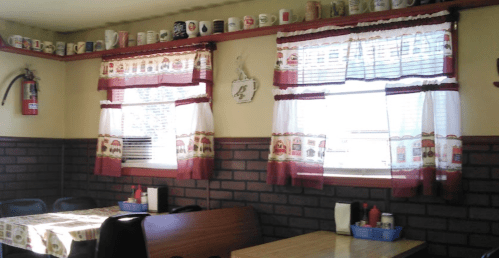 Cozy diner interior with brick walls, checkered tablecloths, and decorative curtains featuring mugs and condiments on tables.