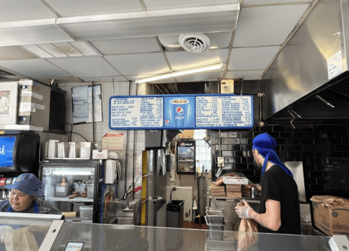 A busy kitchen counter with a menu board, a worker in a blue hat, and a woman preparing food in the background.