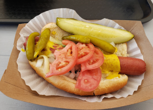 A hot dog in a bun topped with mustard, sliced tomatoes, pickles, and green peppers, served in a paper tray.