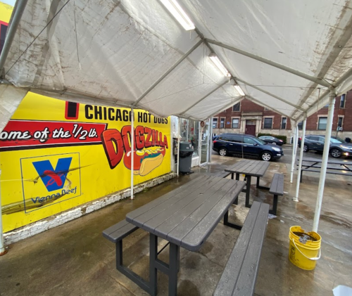 Outdoor seating area under a tent, with picnic tables and a colorful sign for "Dogzilla" hot dogs in the background.