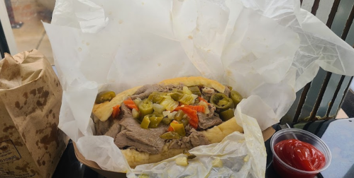 A sandwich filled with sliced beef, jalapeños, and vegetables, served with a side of ketchup in a takeout container.
