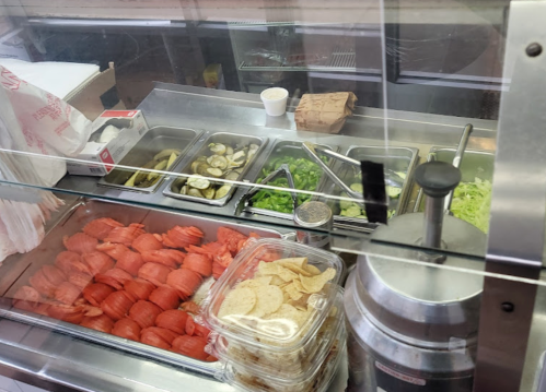 A deli counter displaying sliced tomatoes, pickles, lettuce, and chips in containers, with a drink dispenser in the background.