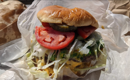 A close-up of a juicy burger with lettuce, tomato, pickles, and melted cheese, wrapped in paper.