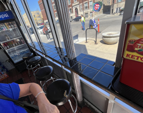 A view from inside a shop showing black stools, a counter, and a street scene outside with pedestrians and a sign.