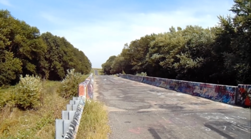A deserted, graffiti-covered bridge stretches into the distance, surrounded by lush greenery and a clear blue sky.