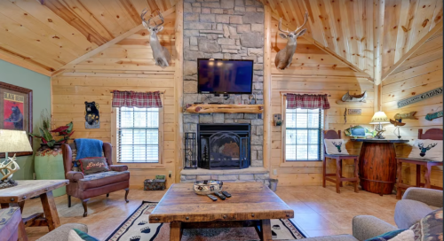 Cozy cabin interior with wooden walls, stone fireplace, mounted deer heads, and rustic furniture.