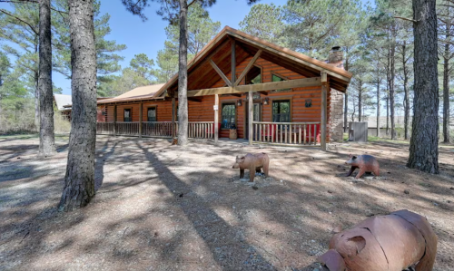 A rustic log cabin surrounded by trees, featuring a porch and decorative bear sculptures in the foreground.