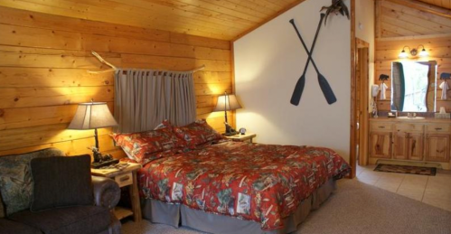 Cozy cabin bedroom with wooden walls, a patterned bedspread, and rustic decor, featuring a bathroom area.