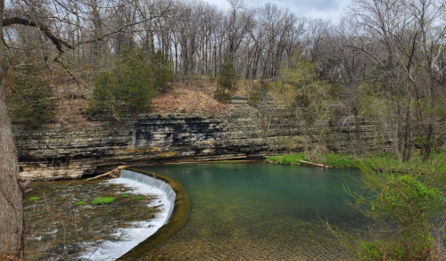 A serene river flows past a rocky cliff, surrounded by trees and greenery under a cloudy sky.