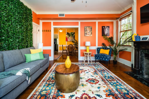 Cozy living room with orange walls, green plants, a gray sofa, colorful rug, and a wooden coffee table.