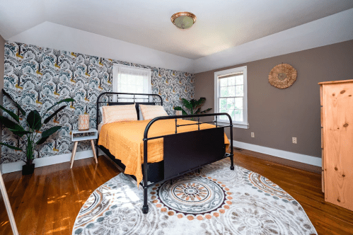Cozy bedroom with a black metal bed, orange bedding, patterned wallpaper, and wooden furniture. Bright and inviting space.