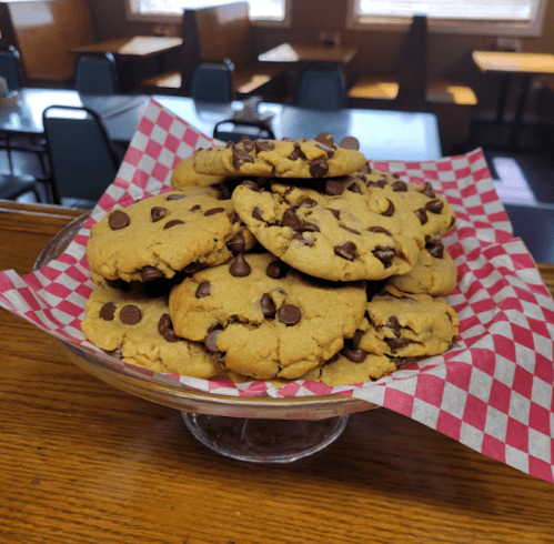 A glass dish filled with chocolate chip cookies on a red and white checkered paper, set in a cozy café.