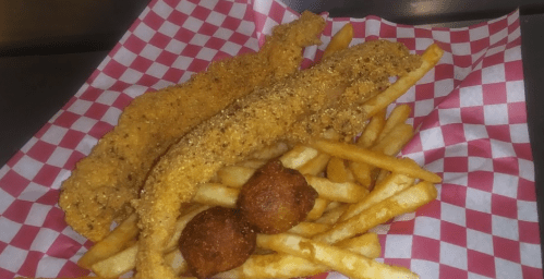 Two crispy fried fish fillets on a bed of golden French fries, with two hushpuppies, served in a red-checkered basket.
