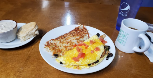 A plate with an omelet topped with cheese and tomatoes, hash browns, a biscuit, and a cup of coffee on a wooden table.