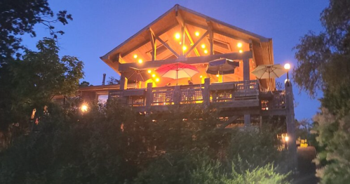A cozy wooden house with a deck, illuminated by warm lights, surrounded by greenery at dusk.