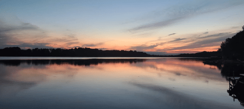 A serene lake at sunset, with colorful clouds reflecting on the calm water and silhouettes of trees in the background.