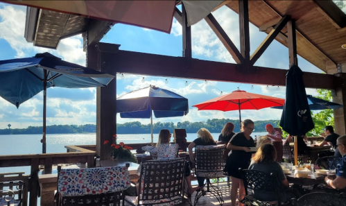 A lakeside restaurant patio with umbrellas, diners enjoying meals, and a scenic view of the water and sky.