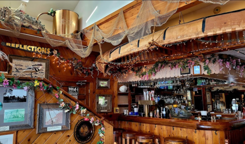 A cozy bar interior with wooden decor, fishing nets, and floral accents, featuring a boat overhead and a well-stocked bar.