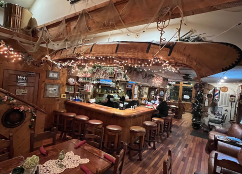 Cozy restaurant interior with a wooden bar, canoe overhead, and warm lighting, featuring wooden tables and chairs.