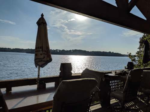 A serene lakeside view from a wooden deck, featuring a sunlit water surface and a patio umbrella.