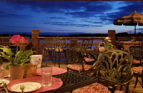 A cozy outdoor dining area with tables and chairs, overlooking a serene lake at sunset, with a pink flower centerpiece.