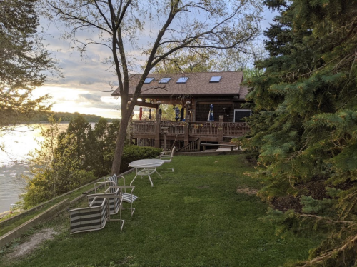 A lakeside cabin with a deck, surrounded by trees and lawn chairs, under a cloudy sky at sunset.
