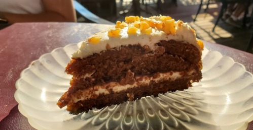 A slice of chocolate cake with cream frosting and chopped nuts, served on a decorative plate.