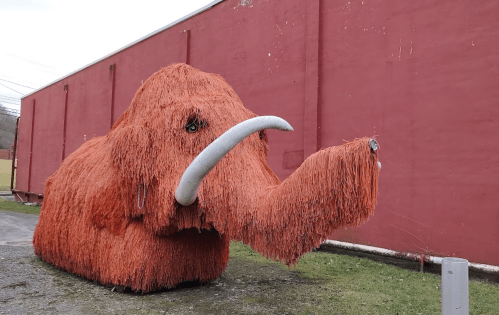 A large straw sculpture of a mammoth with tusks, set against a red wall and grassy area.
