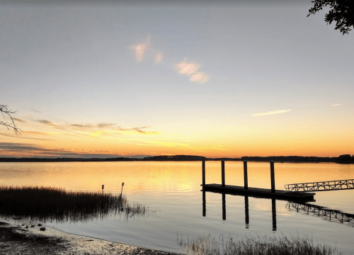 Sunset over a calm lake, with a dock extending into the water and silhouettes of grass along the shore.
