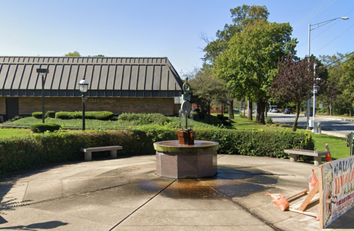 A small fountain surrounded by benches and greenery, with a building in the background and a sign nearby.