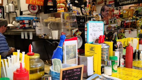 A colorful diner counter with condiments, a cash register, and a display of hot dogs and fries in a vibrant setting.