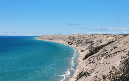 A scenic view of a coastline with sandy cliffs, blue ocean, and clear skies. Waves gently lap at the shore.