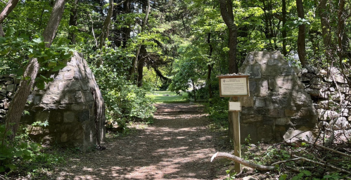 Stone gate entrance to a wooded path, surrounded by lush greenery and a signpost nearby.