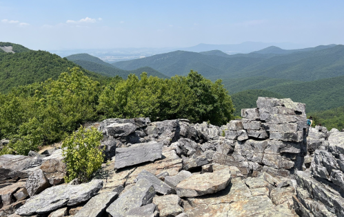 A rocky mountain summit with lush green hills and distant mountains under a clear blue sky.