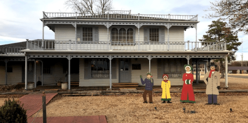 A two-story house with a porch, featuring cutout figures in front, set against a cloudy sky.