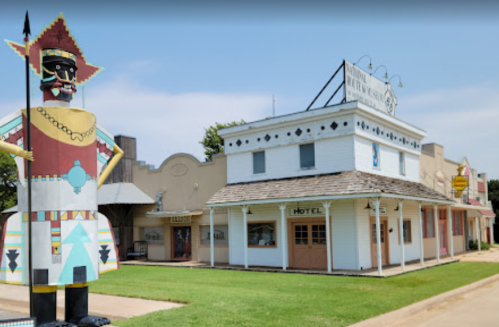 A colorful roadside attraction featuring a large wooden figure and a vintage-style hotel building.