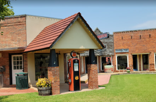 A vintage gas station with a red roof and classic gas pump, surrounded by brick buildings and green grass.