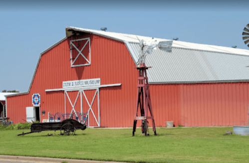Red barn-style building with a sign reading "Farm & Ranch Museum," featuring a windmill and vintage wagon in front.