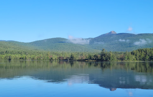 A serene lake surrounded by lush green mountains under a clear blue sky, with reflections on the water's surface.