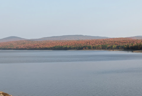 A serene lake surrounded by autumn-colored trees and distant hills under a clear blue sky.